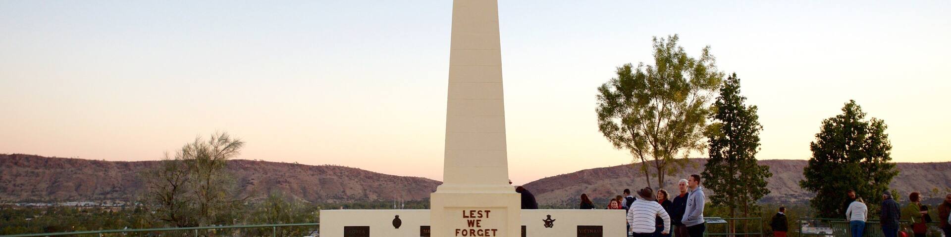 A tall white obelisk with 'Lest We Forget' inscribed at its base, surrounded by a chain-link fence, set against a backdrop of hills and trees.
