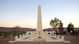 Et højt, hvidt obelisk med teksten 'Lest We Forget' indgraveret ved foden, omgivet af et pigtrådshegn, foran en bakke og træer.