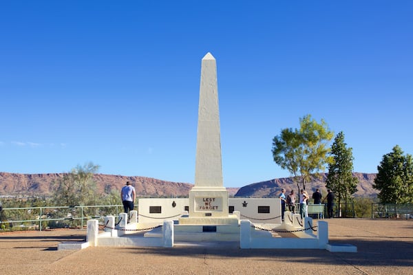 Ein hohes, weißes Obelisken-Denkmal mit der Inschrift "Lest We Forget" in einem Park mit blauem Himmel und roten Felsen im Hintergrund.