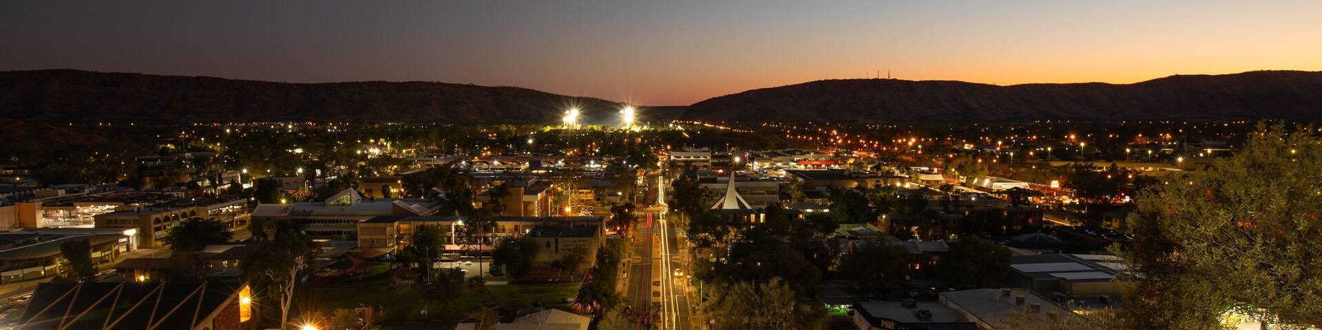 A town at dusk with illuminated streets and buildings, surrounded by hills.