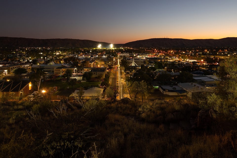 Uma cidade ao anoitecer, com ruas e edifícios iluminados, cercada por colinas.