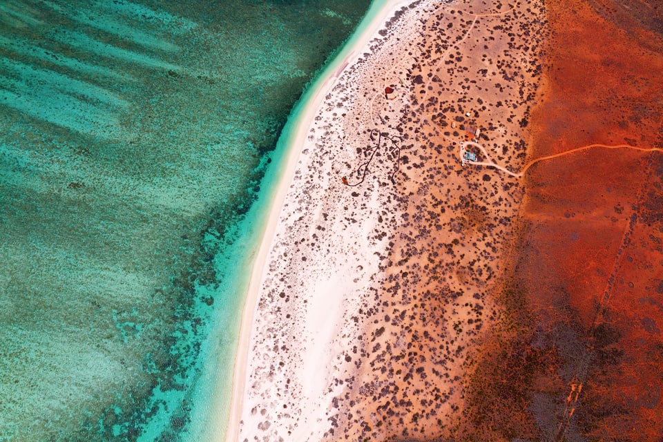 Vista aérea de uma praia com um degradê de cores marcante, do azul-turquesa para o vermelho, e pedras espalhadas.
