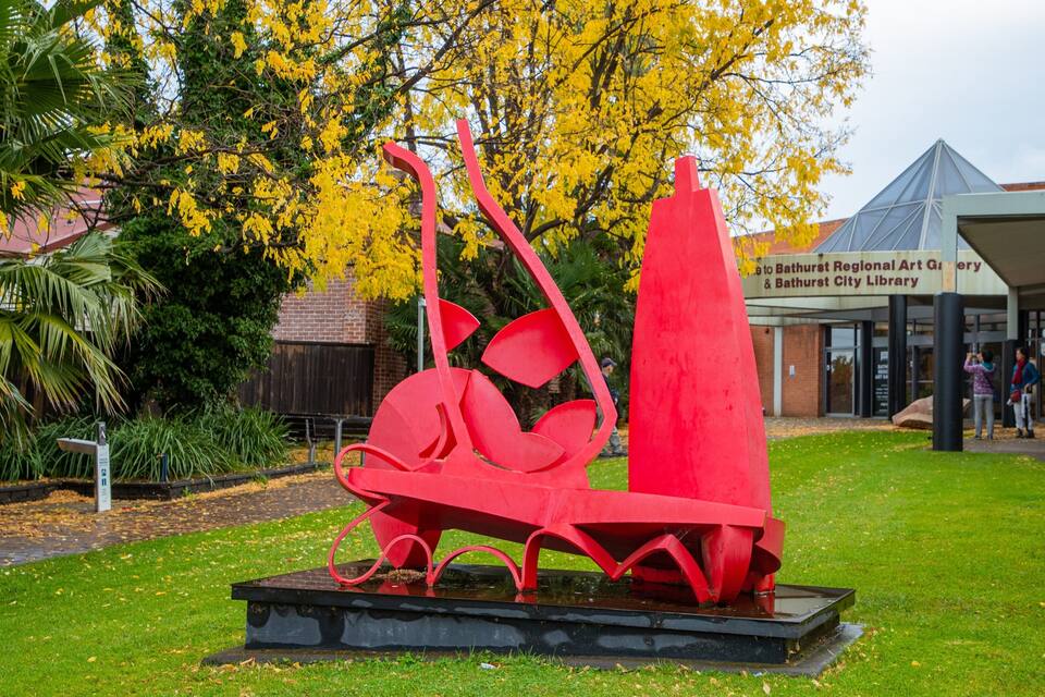A red abstract sculpture in a park with a building labeled "Bathurst Regional Art Gallery & Bathurst City Library" in the background.