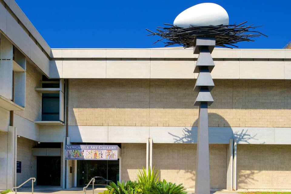 A modern building with a distinctive sculpture on top, a sign for Newcastle Art Gallery, and a clear blue sky.