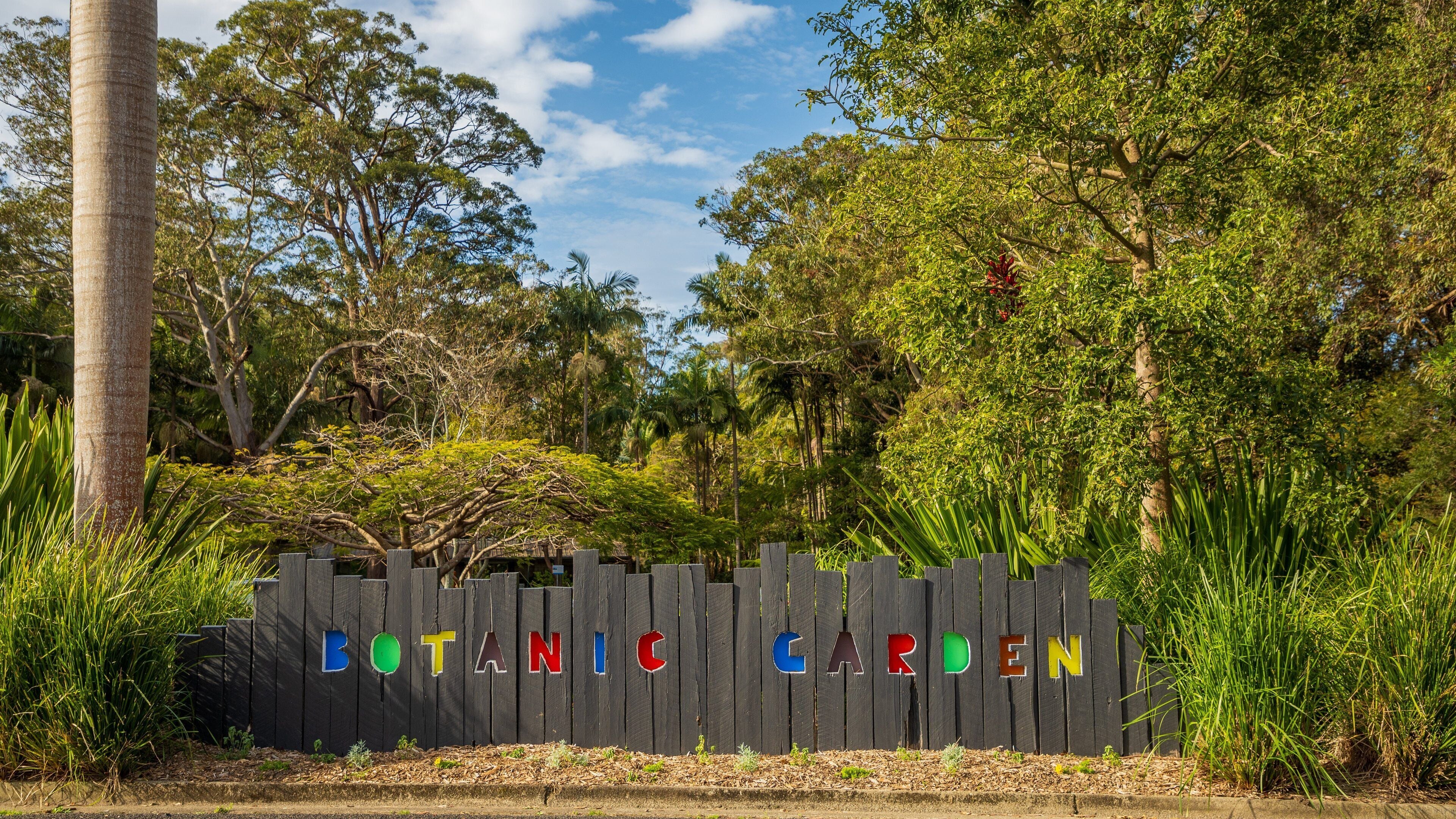 A wooden fence with colorful letters spelling "BOTANIC GARDEN" amidst lush greenery and trees.