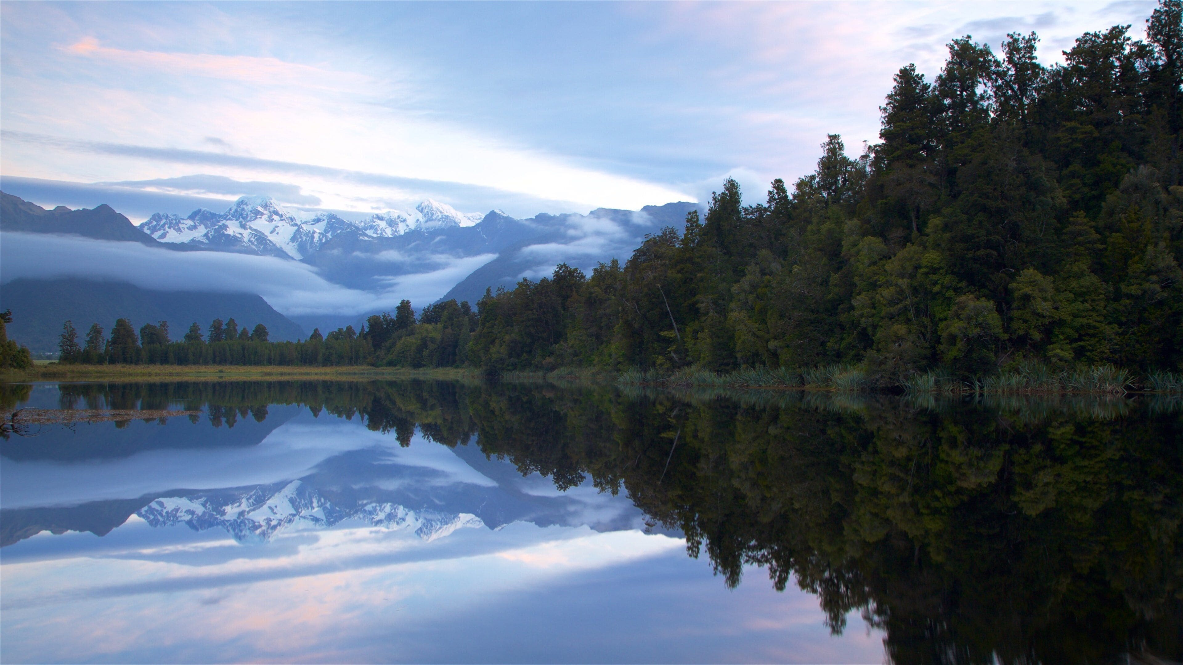 A calm lake reflecting snow-capped mountains and a dense forest.