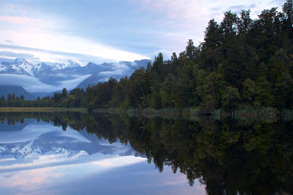 A calm lake reflecting snow-capped mountains and a dense forest.