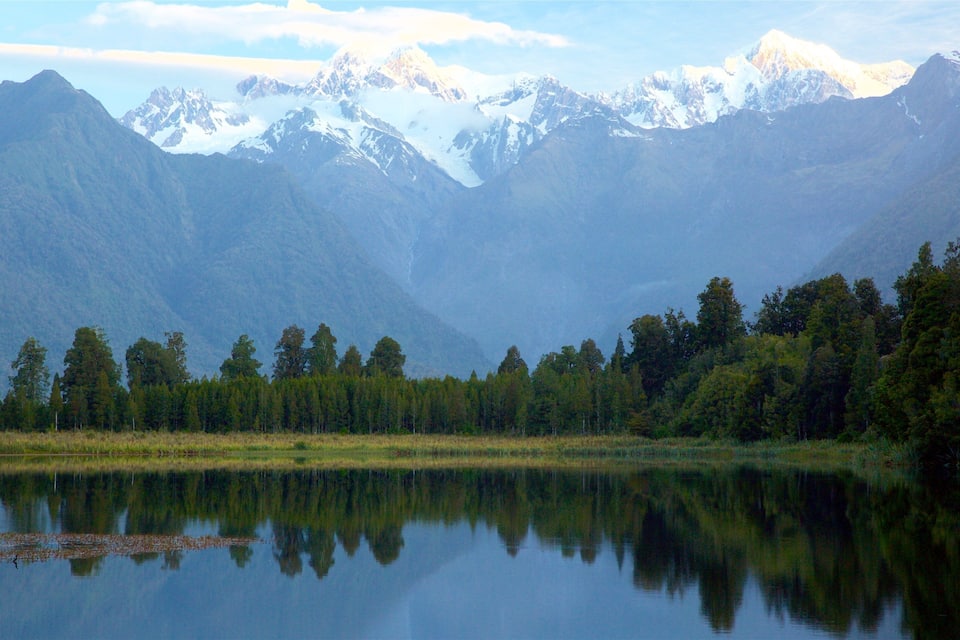 A calm lake reflecting trees and mountains with snow-capped peaks in the background.