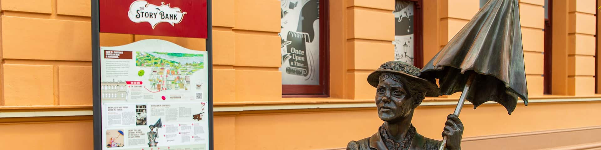 Une statue représentant une femme tenant un parapluie, un banc rouge et une pancarte indiquant « Banque à histoires ».