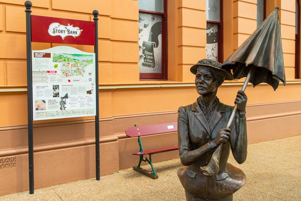 A statue of a woman holding an umbrella, a red bench, and a 'Story Bank' sign.