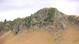 A rugged mountain with a cross on top, rocky cliffs, and sparse vegetation.