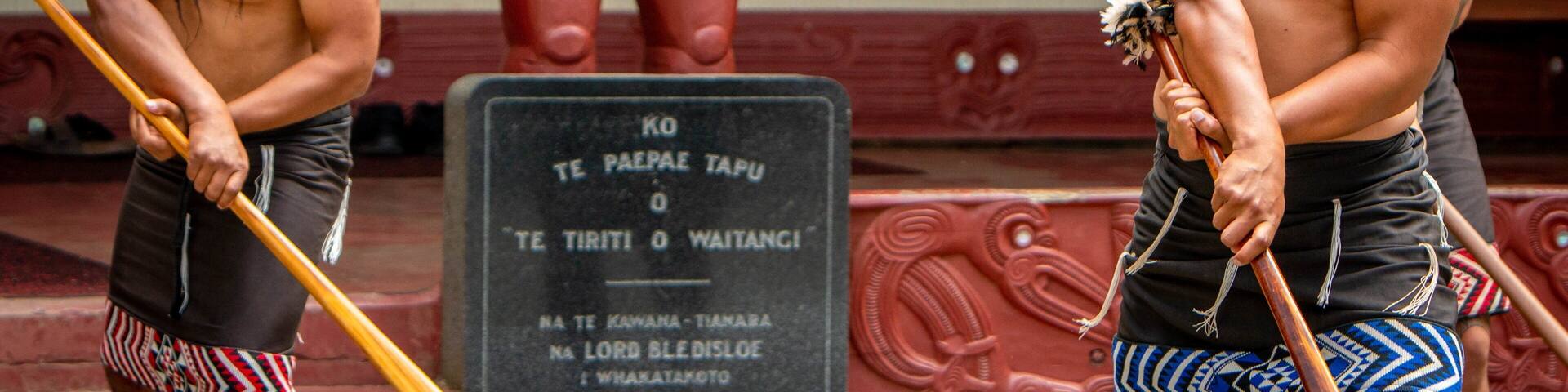 Deux personnes tenant des armes traditionnelles, une plaque de pierre portant des inscriptions et une statue en arrière-plan.
