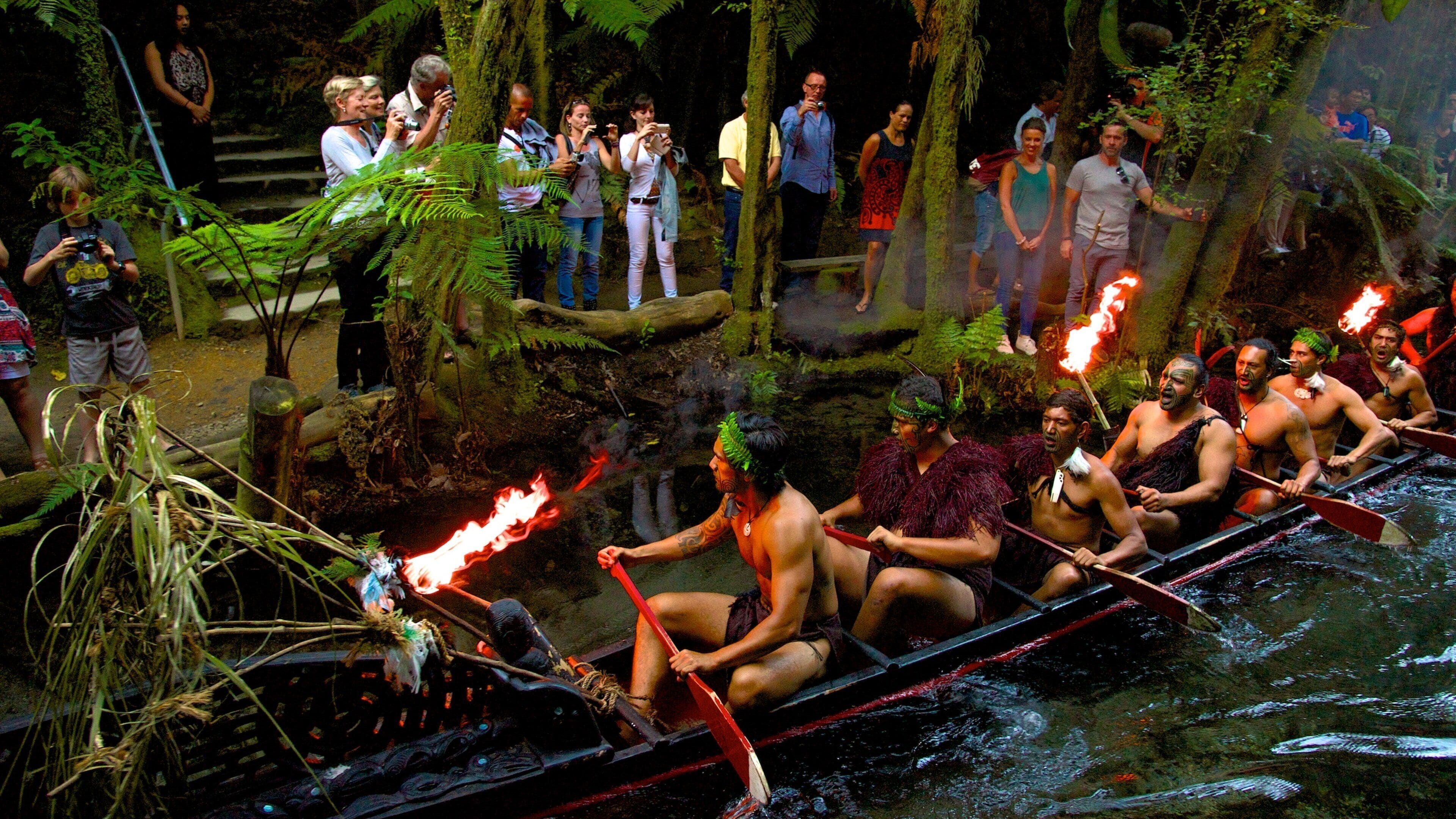 Una carrera de canoa con participantes vestidos con atuendos tradicionales, en un entorno boscoso con espectadores.
