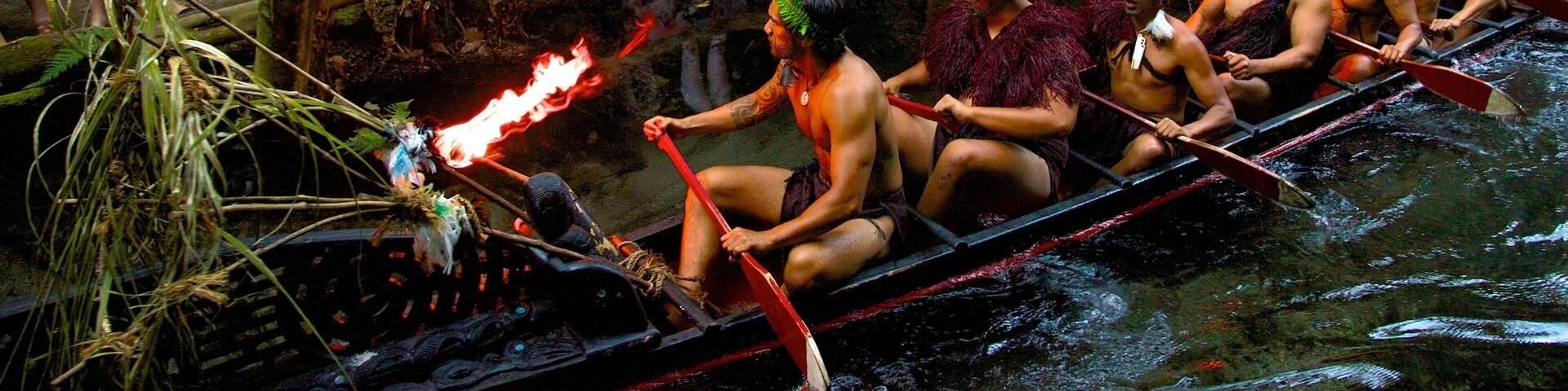 A canoe race with participants in traditional attire, surrounded by a forested area with spectators watching.