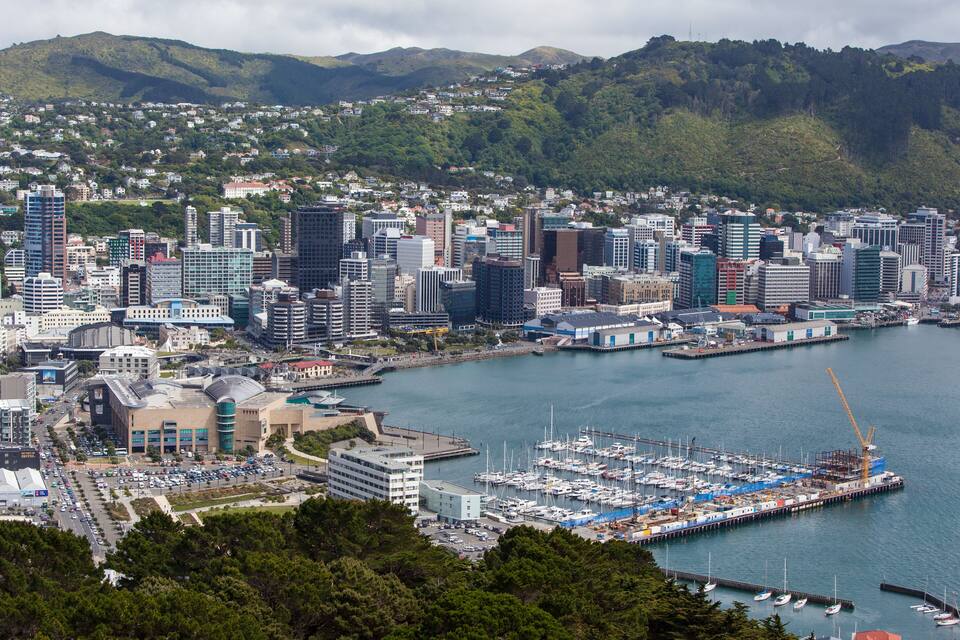 A coastal cityscape with a marina, modern buildings, and a mountainous backdrop.