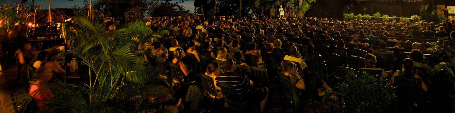Un cine al aire libre de gran tamaño con una multitud de personas sentadas en filas frente a una pantalla grande.