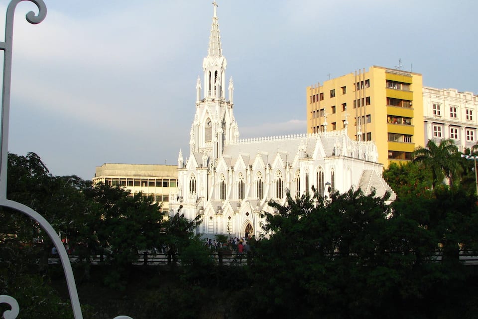 En vit katedral med ett högt torn, omgiven av träd och moderna byggnader.