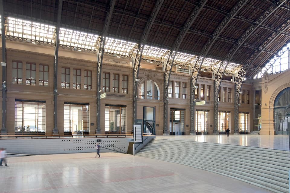 Una estación de tren espaciosa y bien iluminada, con techos altos, amplias ventanas y varias escaleras.