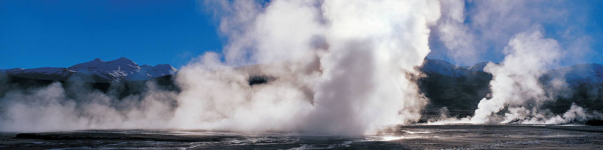 A geyser erupting with steam against a backdrop of snow-capped mountains.