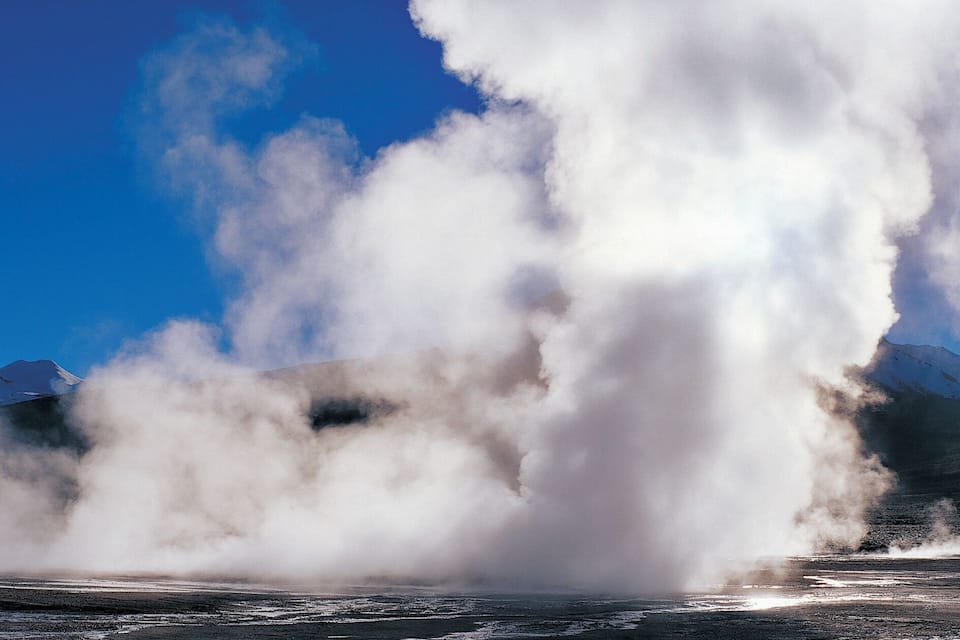 Geysir purkautuu, ja höyryä nousee ilmaan lumihuippuisten vuorten taustaa vasten.