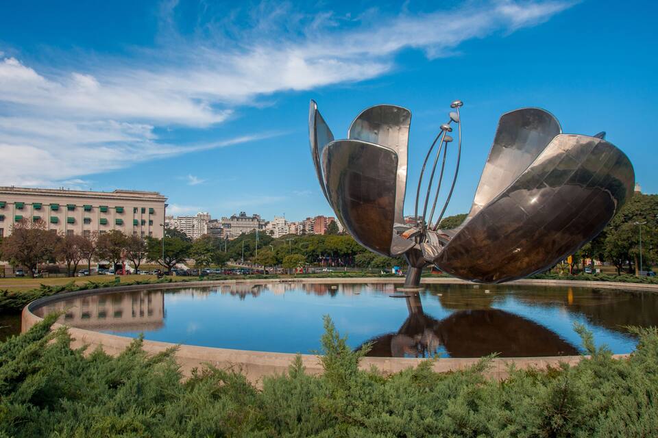 A metallic sculpture in a park with a reflective pond, surrounded by greenery and buildings in the background.