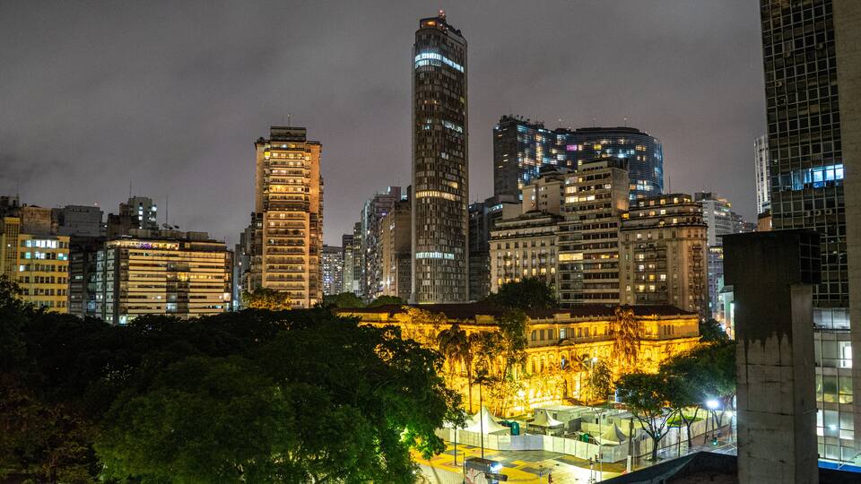 Paisagem urbana noturna com edifícios iluminados e uma praça central com árvores.