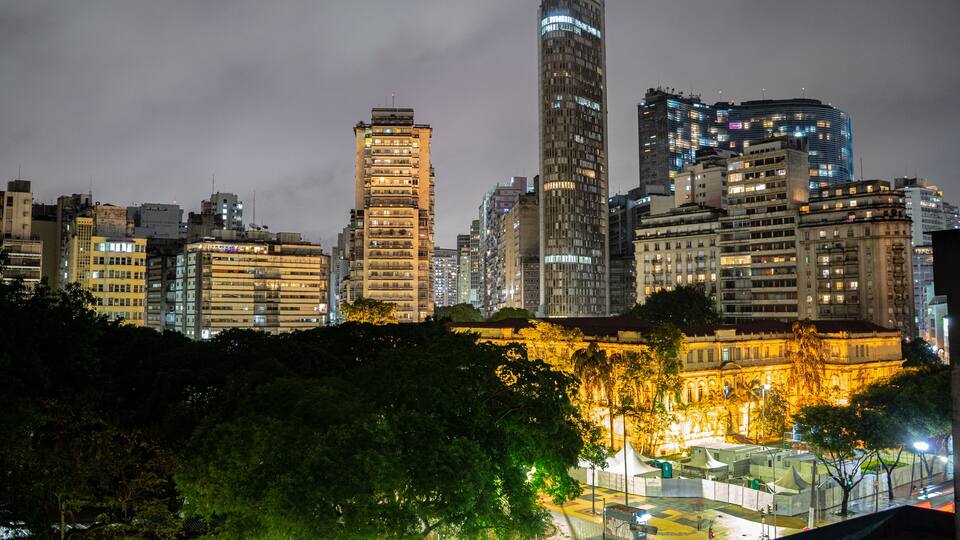 Um horizonte urbano noturno com edifícios iluminados, um parque com árvores e um edifício histórico grande em primeiro plano.