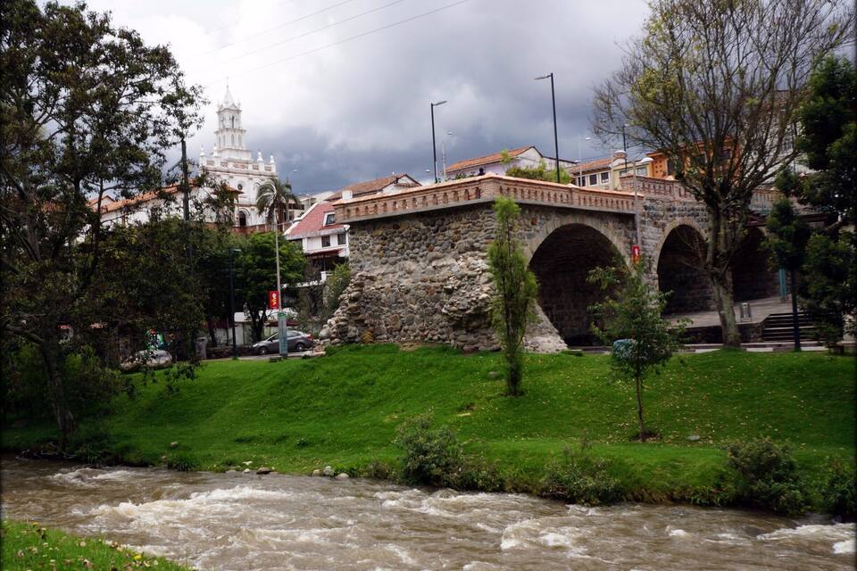 A stone bridge over a river, a white church with a bell tower, and lush greenery.
