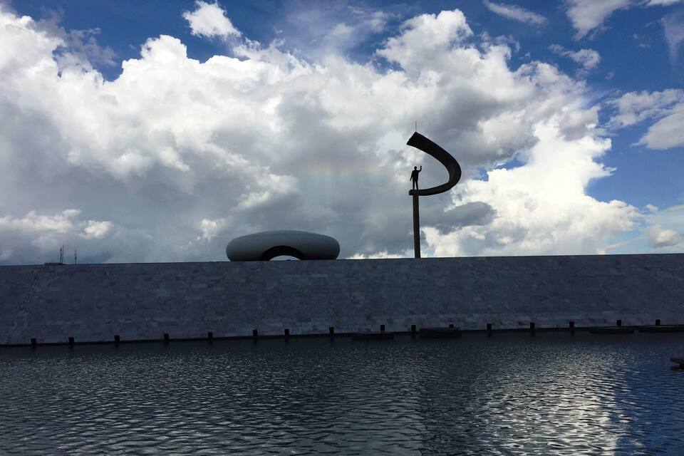 A sculpture of a person standing on a curved structure, a large circular building, and a body of water under a cloudy sky.