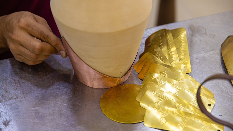Una persona trabaja con un objeto cilíndrico grande color beige y hojas texturizadas de color dorado sobre una mesa metálica.