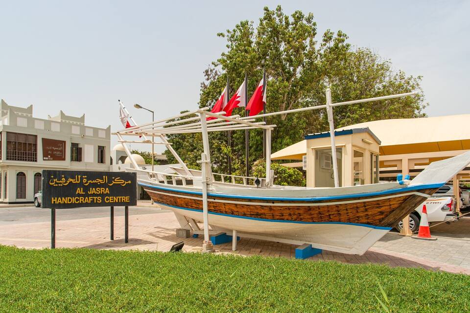 Ein traditionelles hölzernes Boot mit roten Flaggen, vor dem Al Jasra Handicrafts Centre geparkt.