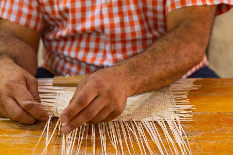 Uma pessoa trançando palha sobre uma mesa de madeira.