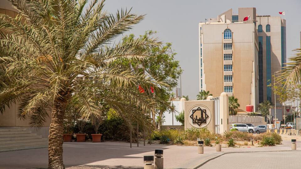 A palm tree-lined street with a tall building featuring a prominent emblem and flags.
