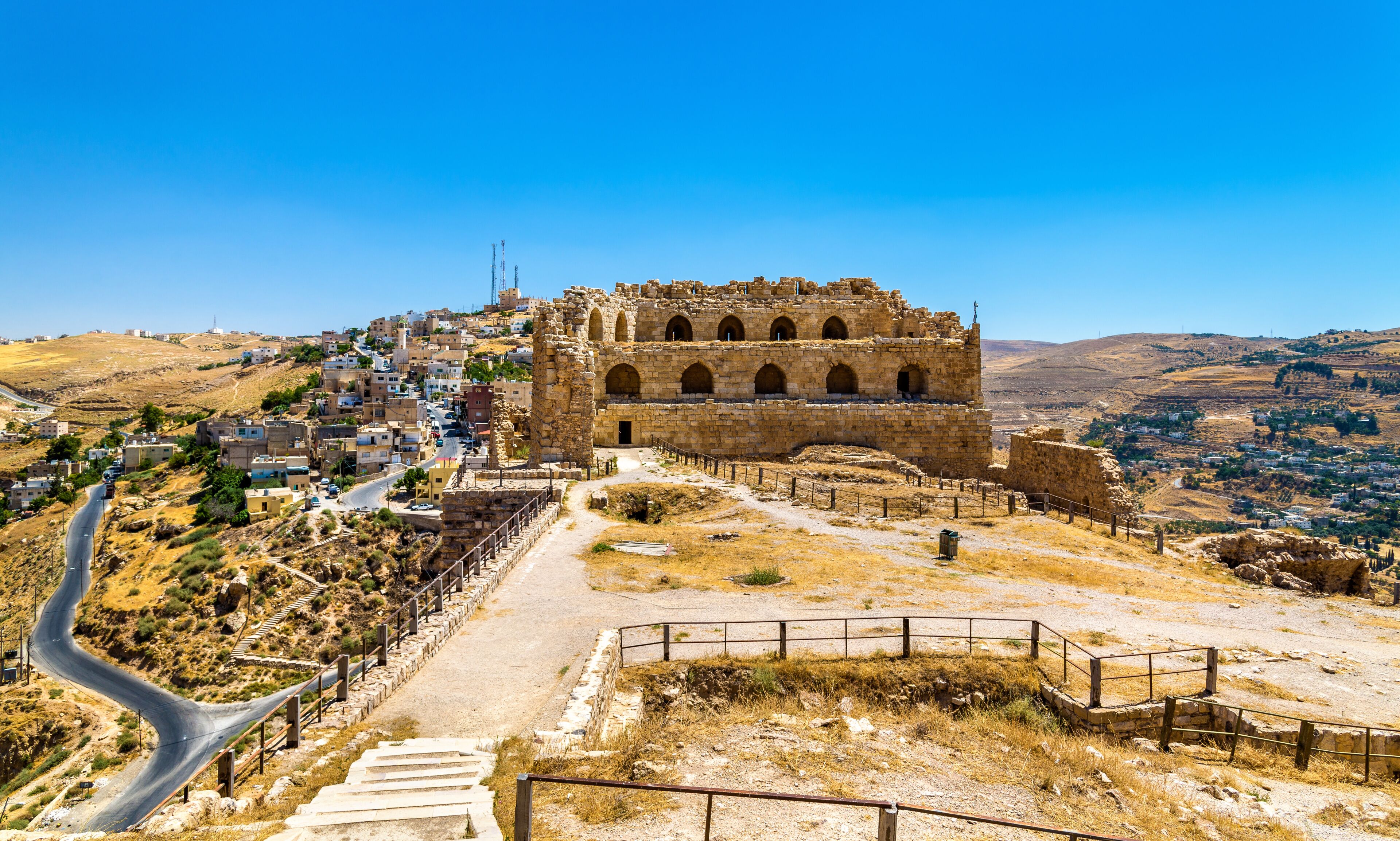 A historic stone fortress with arched windows, a winding road, and a hillside town in the background.