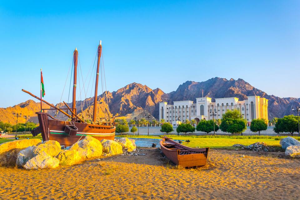 A coastal scene with a large ship, smaller boats, and a grand building with a mountainous backdrop.