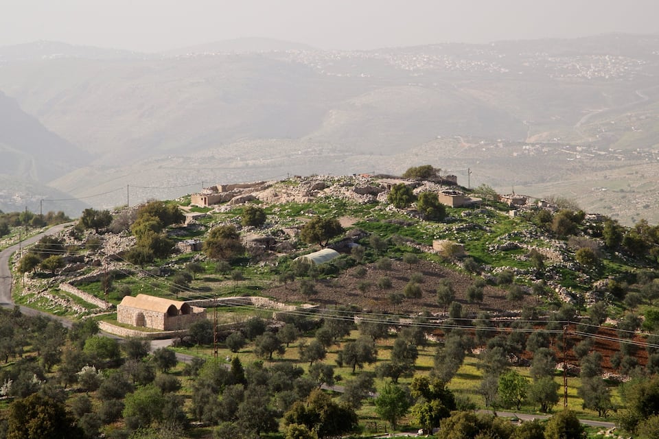 A mountainous landscape with a winding road, scattered buildings, and olive groves.
