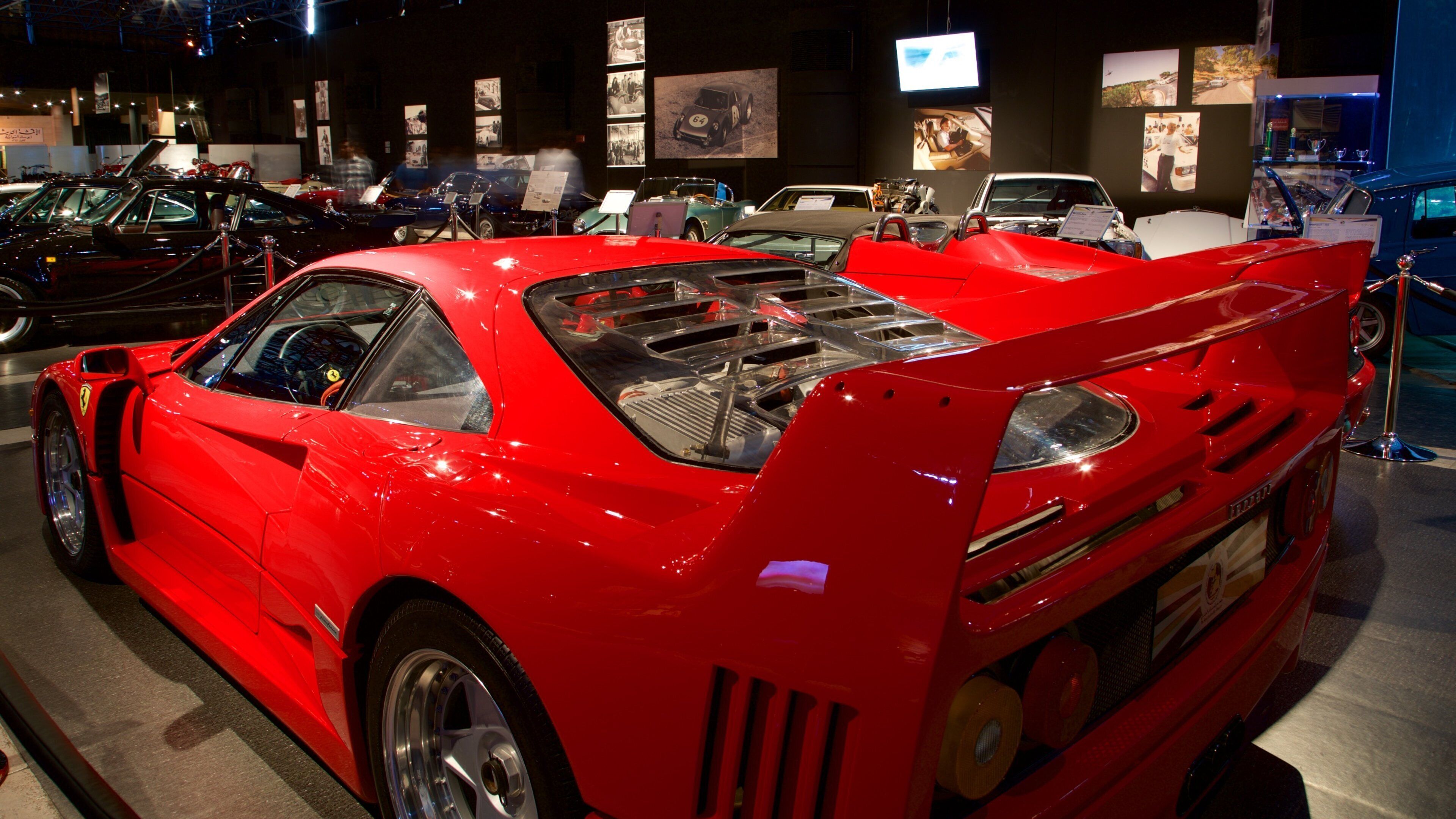 A red sports car with a rear spoiler and visible engine components, displayed at an indoor exhibition.