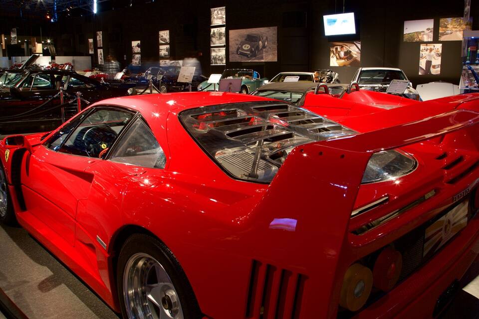 A red sports car with a rear spoiler and visible engine components, displayed at an indoor exhibition.