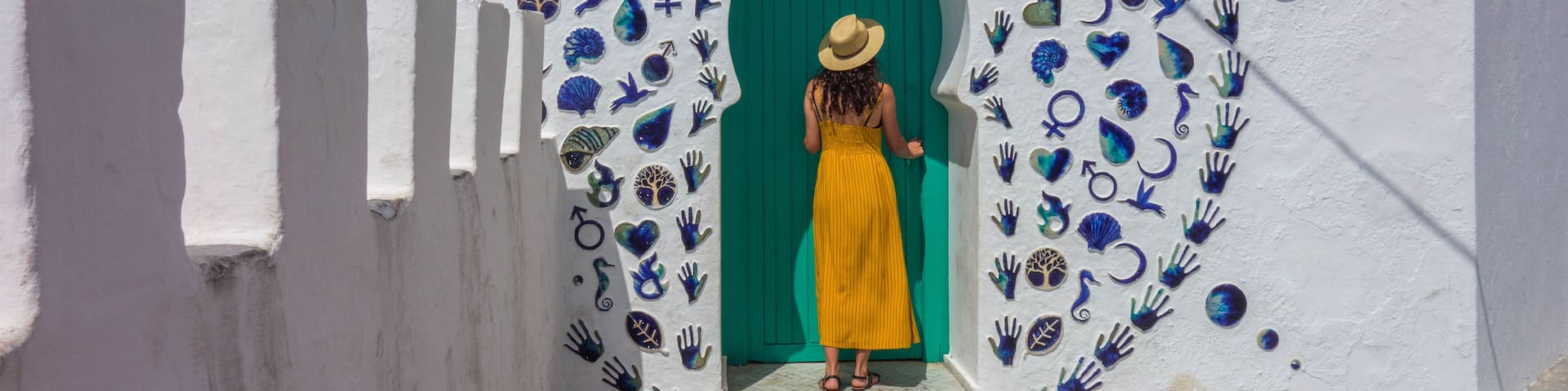 A woman in a yellow dress standing at a green door with decorative handprints and symbols on the wall.