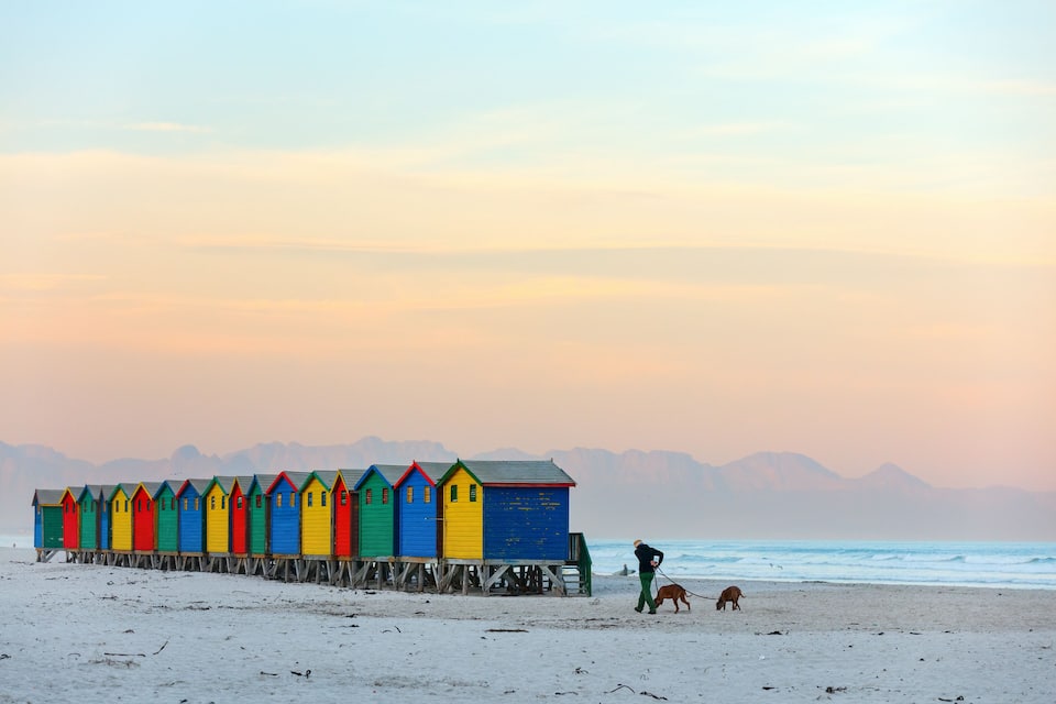 A row of colorful beach huts on stilts along a sandy beach with a person walking a dog and mountains in the background.