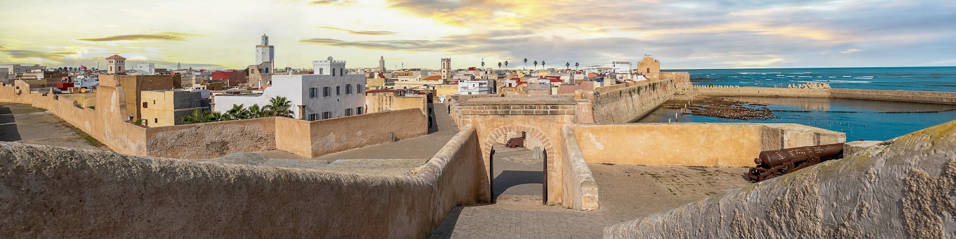 Un paisaje urbano costero con una fortificación histórica, cielo despejado y mar tranquilo.