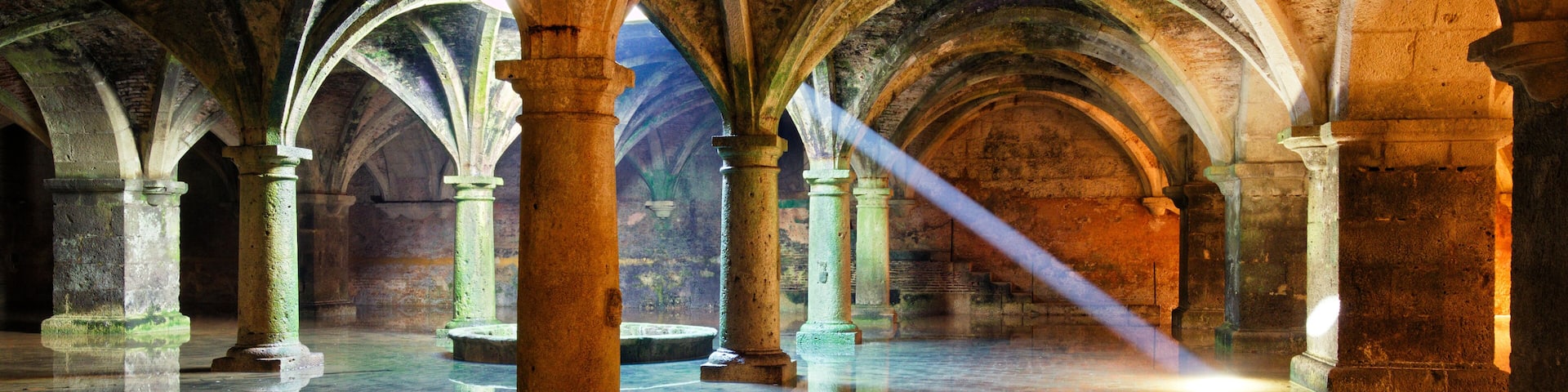 An interior with stone arches and columns, a reflective floor, and a beam of light.