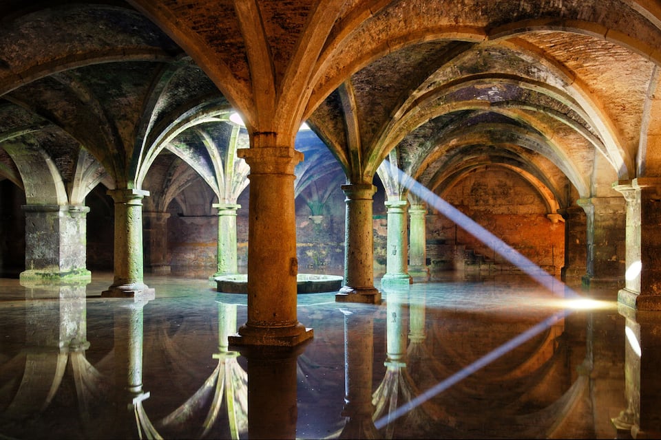 Interno caratterizzato da archi e colonne in pietra, un pavimento riflettente e un raggio di luce.