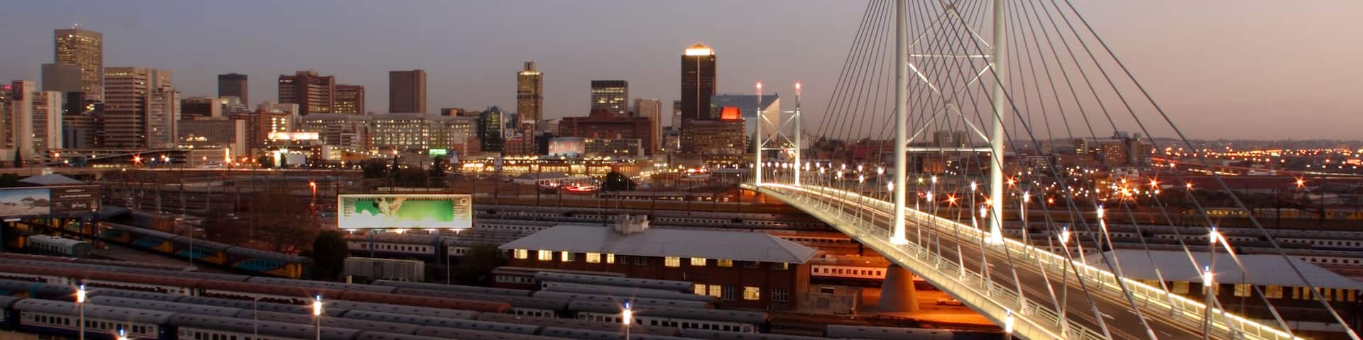 Un horizonte urbano al anochecer con un puente atirantado destacado y varias vías de tren.