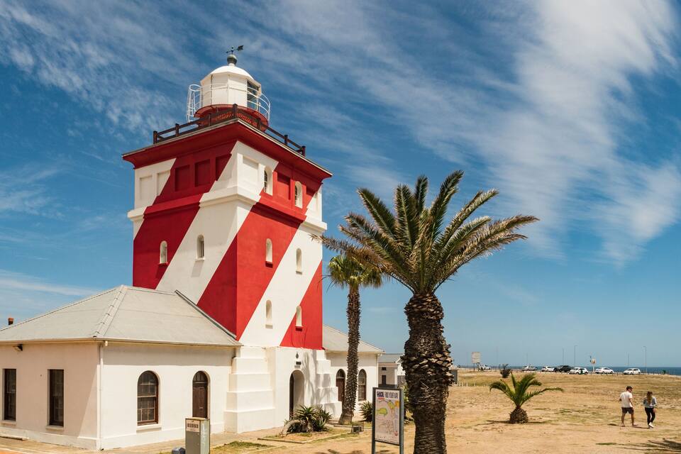Um farol vermelho e branco com um indicador de vento, palmeiras e um céu azul límpido.