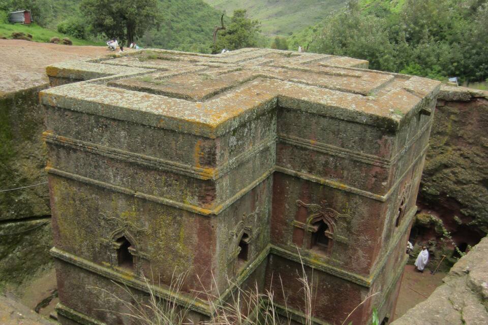 Una estructura de piedra con varios niveles y aberturas arqueadas, rodeada de verdes colinas y un cielo despejado.