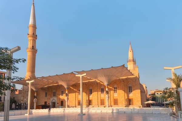 A mosque with a tall minaret and a smaller tower, surrounded by a covered courtyard with white benches.