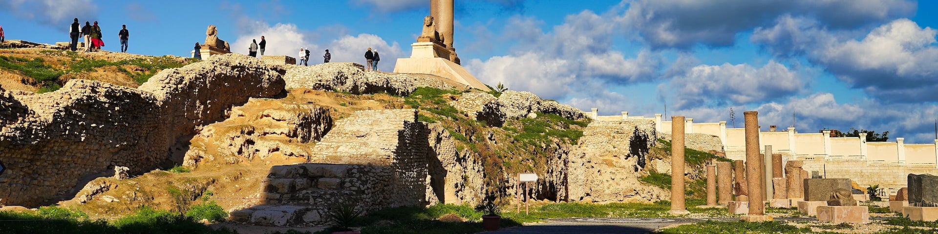 Una columna alta con una estatua en la cima, rodeada de ruinas y un cielo azul despejado.
