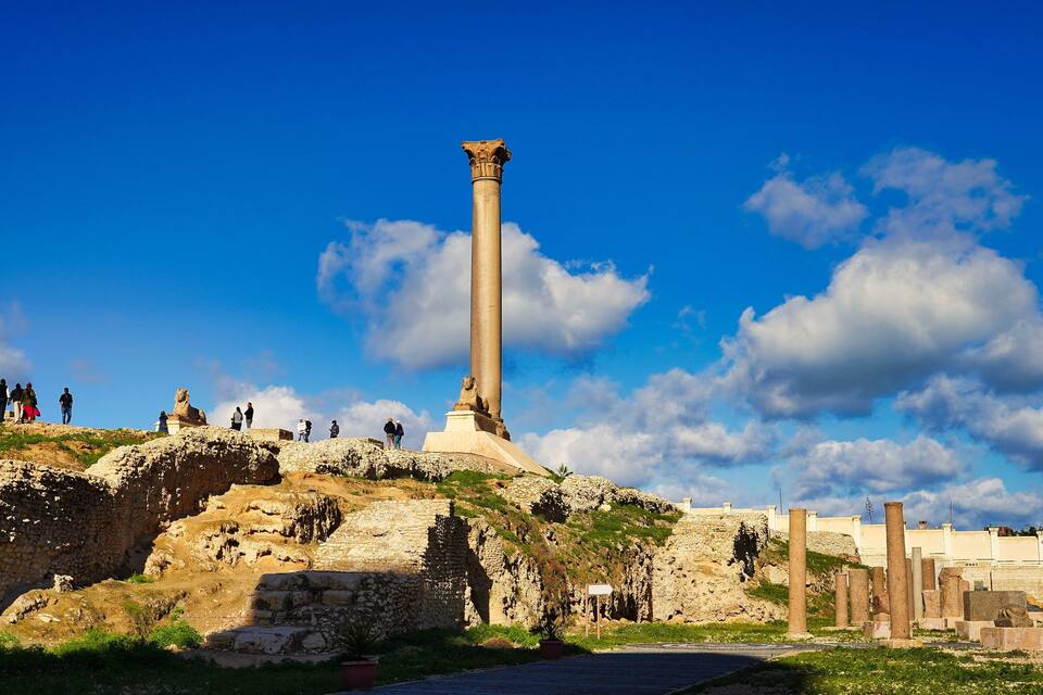 A tall column with a statue on top, surrounded by ruins and a clear blue sky.