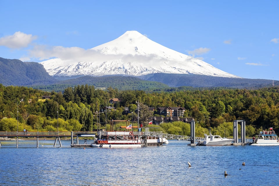 A snow-capped mountain, a bridge, boats, and a forested area.